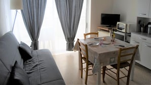 A living room with a sofa, dining table, chairs, a TV, a microwave, and a clock on the wall. - Apartment with Balcony 5 min from Spa - Pets allowed (Balaruc-les-Bains)