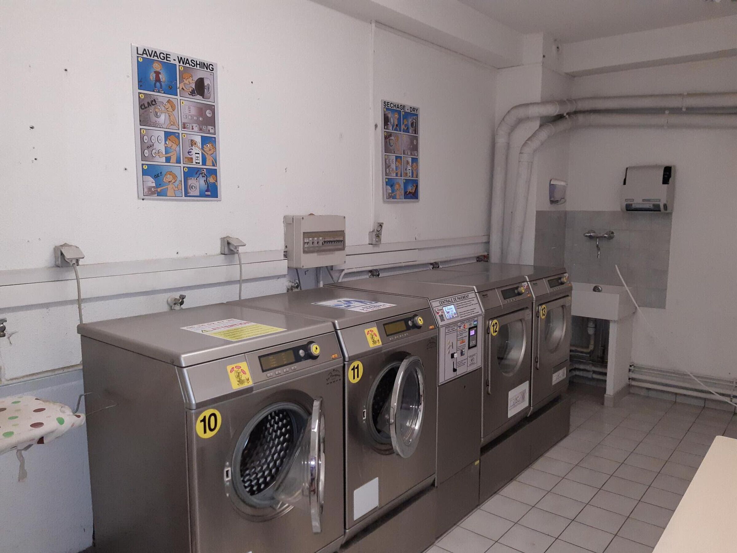 A laundry facility with four washing machines, numbered 10 to 13, and two posters on the wall.