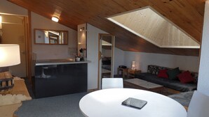 A cozy loft-style bedroom with a sloped ceiling, a small kitchenette, a dining area with a table and chairs, and a sofa with cushions.