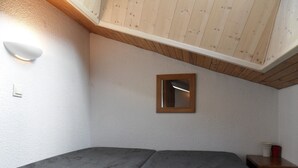 A bedroom with a sloped ceiling, a bed with a grey mattress, a wooden headboard, a small wooden-framed mirror, and a nightstand with a red object.