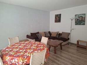 A living room with a sofa, a dining table with a floral tablecloth, and two paintings on the wall.