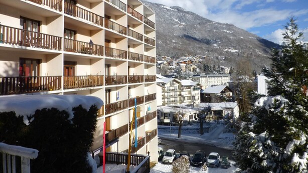 Un hotel de varios pisos con balcones, vista a una montaña nevada y automóviles estacionados en primer plano.