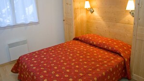 Quarto com cabeceira de madeira, duas luminárias fixadas na parede, cama com colcha florida vermelha e janela com cortinas brancas.