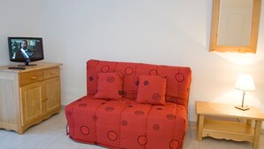 Quarto com sofá vermelho, armário de madeira, televisão, mesinha com abajur e espelho na parede.