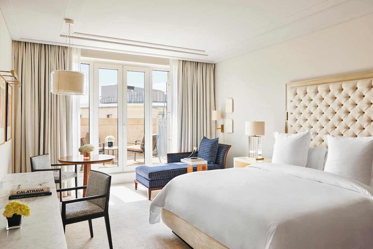 Habitación de hotel con una cama grande, un escritorio con silla, una mesita con otra silla y vistas al exterior a través de las ventanas.