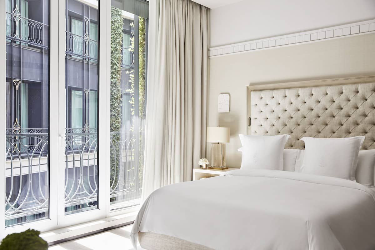 Una habitación de hotel con una cama grande, una mesita de noche con lámpara y vistas a un edificio a través de una ventana con cortinas.