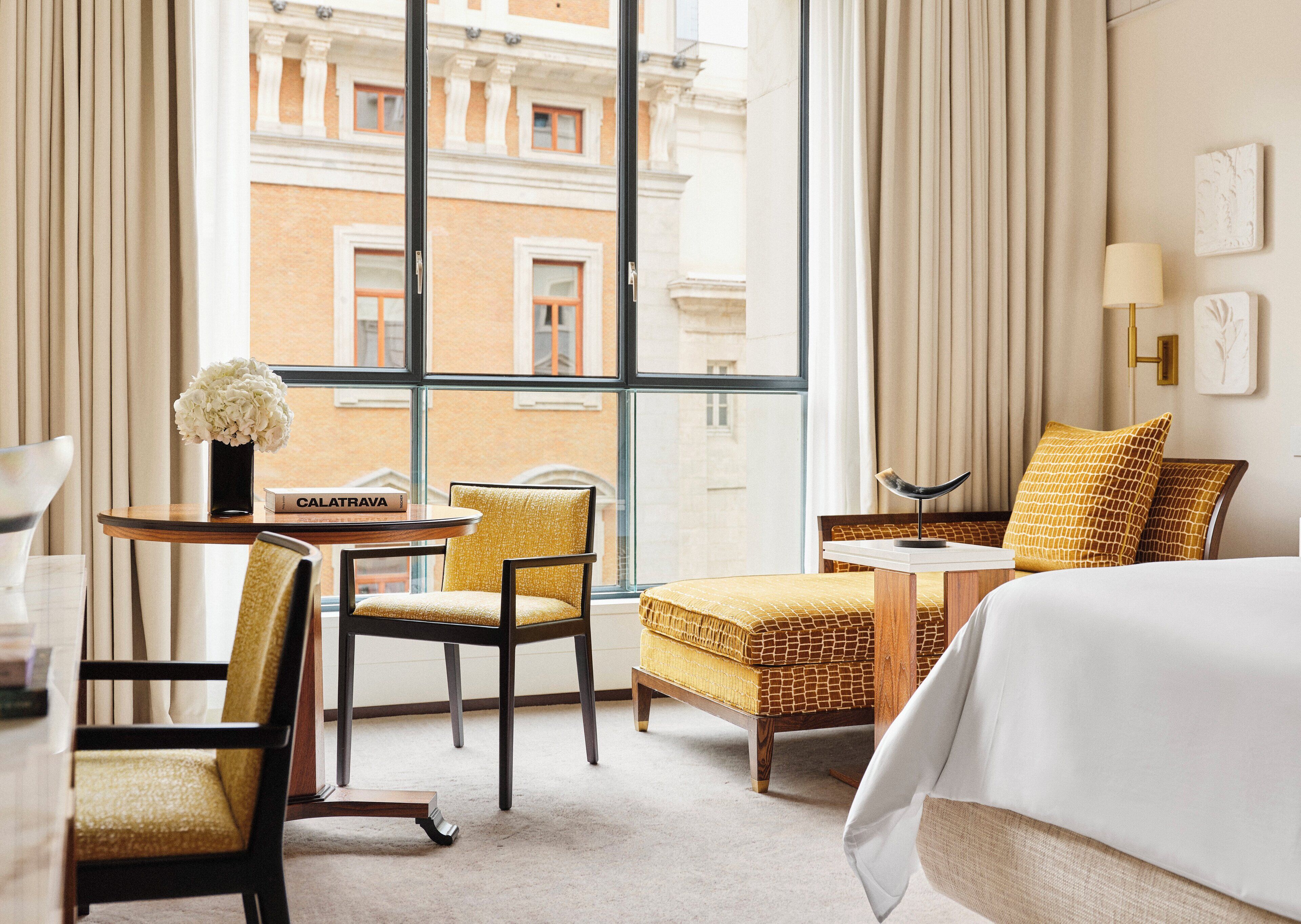 Habitació d'hotel amb un llit, una cadira, una taula petita amb un llibre, un ram de flors i vista d'un edifici a través de grans finestres.