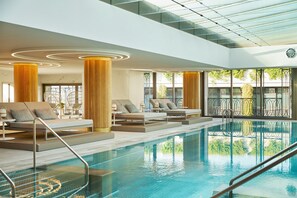 Un área de piscina interior moderna con sillones de descanso, techo de cristal y vistas al exterior.