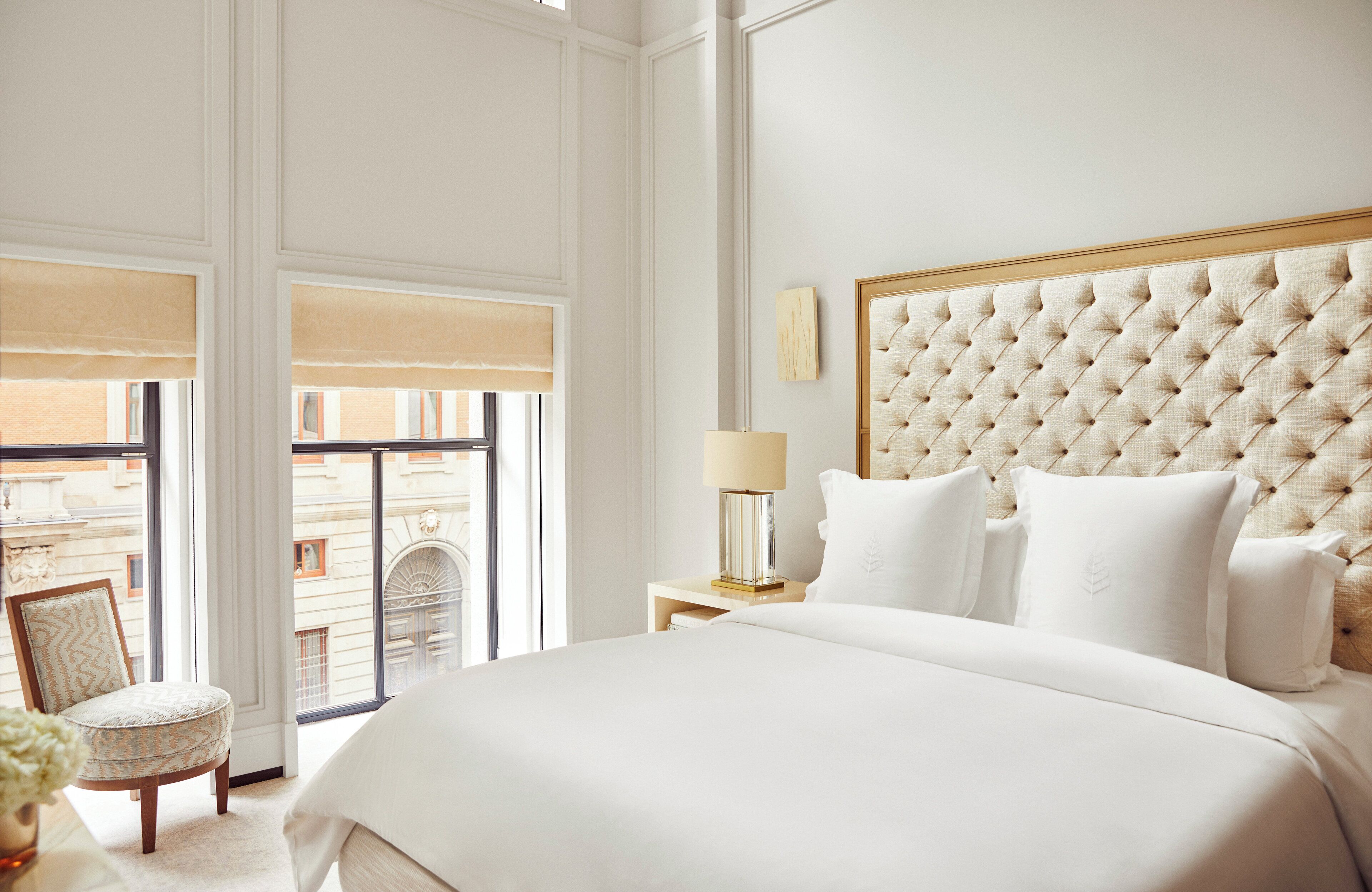 Habitación de hotel con una cama grande, una silla, una mesita de noche con lámpara y vistas a la ciudad a través de amplios ventanales.