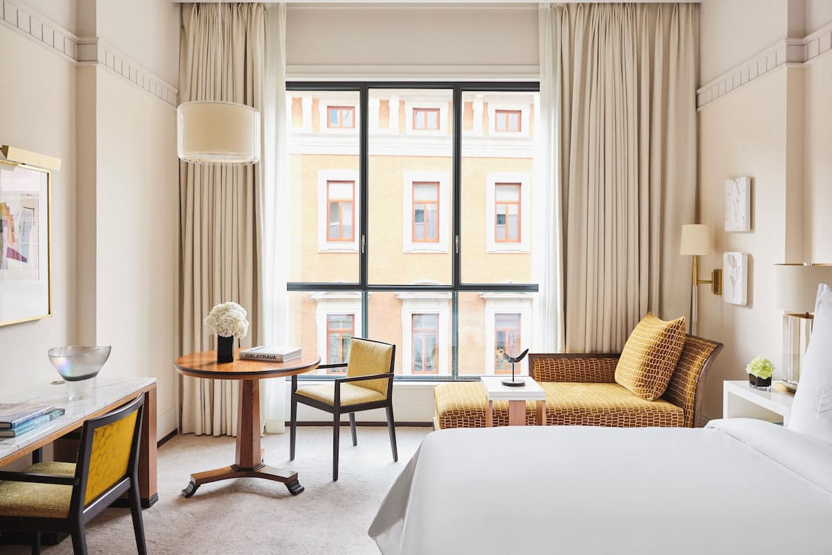 Habitación de hotel con un ventanal grande, una cama, un escritorio con una silla, una mesita con un florero y un sofá con cojines.