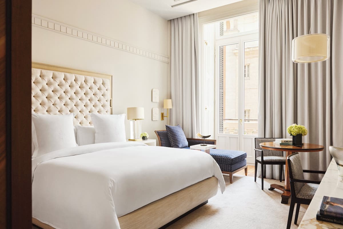 Habitación de hotel con una cama grande, una mesita de noche con lámpara, una silla y vista a la ciudad por la ventana.