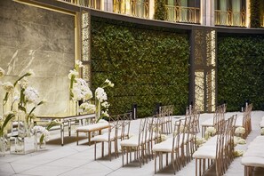 A formal indoor setting with rows of white chairs and tables, a marble wall, and a green leafy wall.