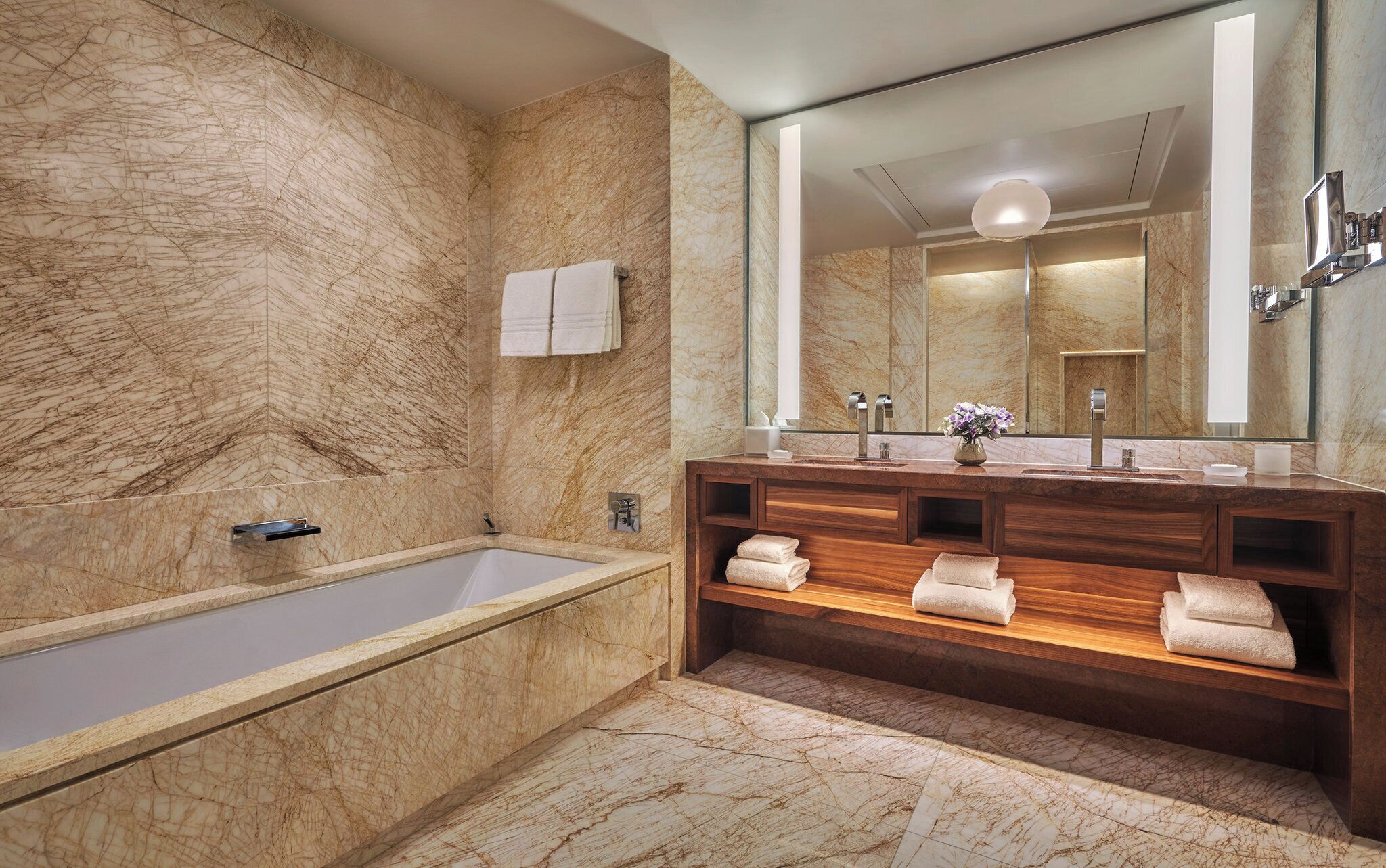 Un baño moderno con pared de mármol, una bañera independiente, un tocador de madera con espejo y estanterías empotradas para toallas.