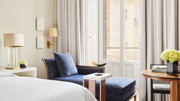 Ein Hotelzimmer mit einem Bett, einem blauen Polsterstuhl, einem kleinen Tisch mit Blumenvase und Blick auf die Stadt durch große Fenster.