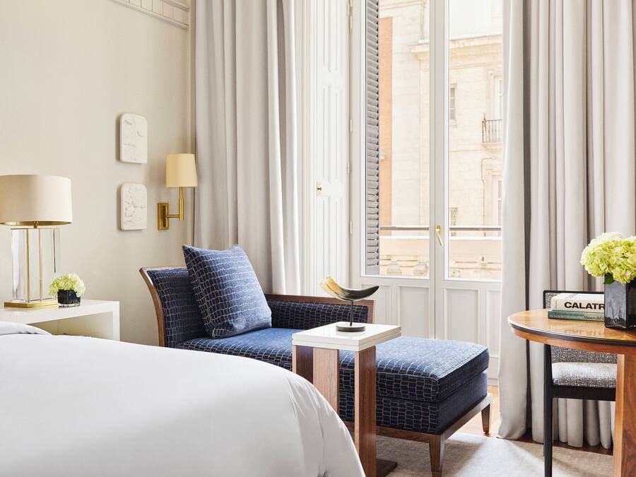 Ein Hotelzimmer mit einem Bett, einem blauen Polsterstuhl, einem kleinen Tisch mit Blumenvase und Blick auf die Stadt durch große Fenster.