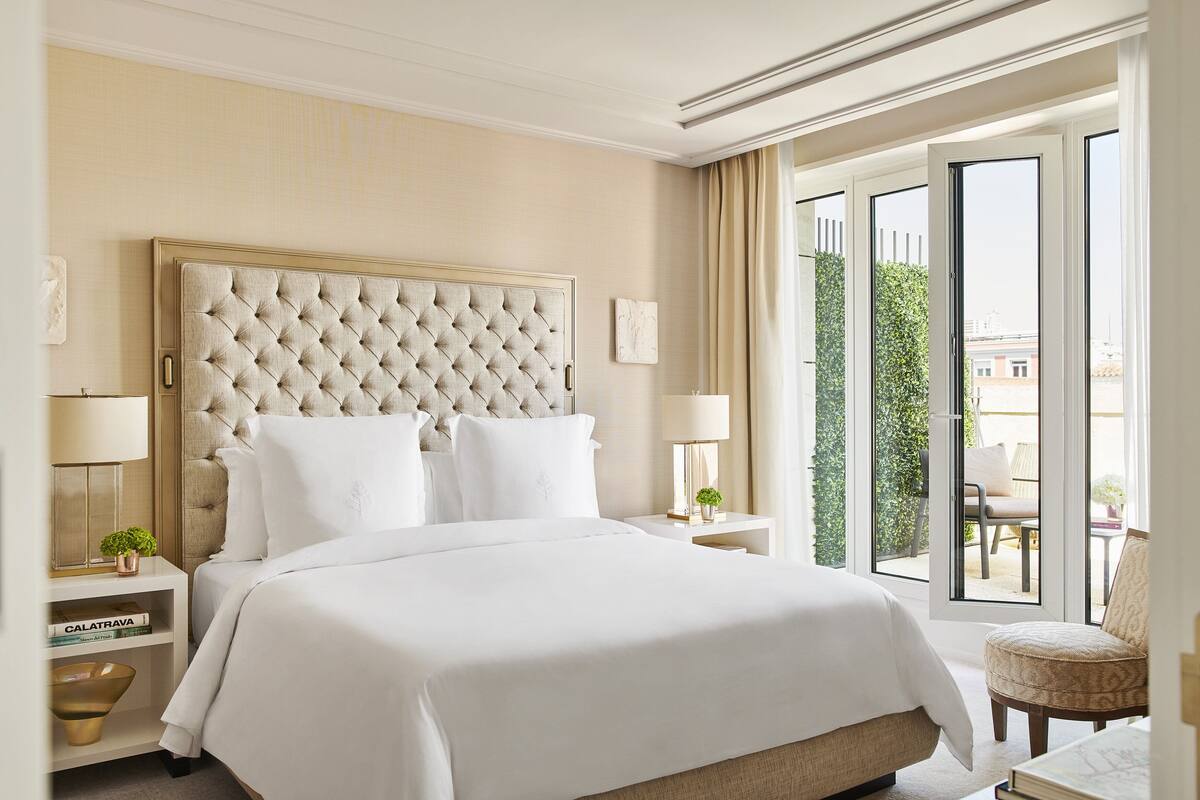 Habitación de hotel con una cama grande, un cabecero capitonado, una mesita de noche con lámpara, una silla y vistas al exterior.