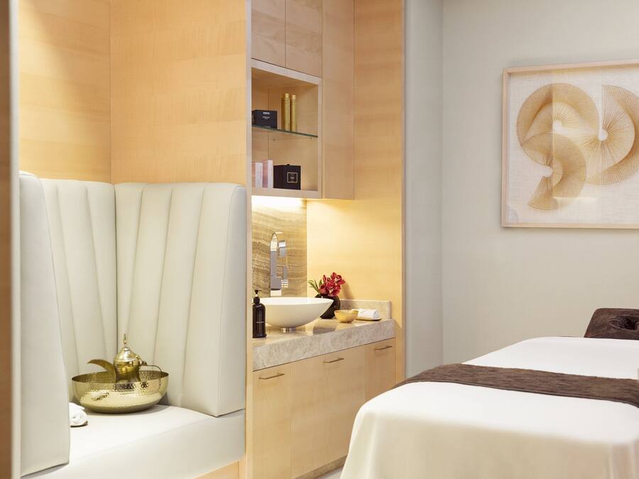 A modern hotel room with a large bed, a sitting area with a white chair, a vanity with a mirror and sink, and a framed artwork on the wall.