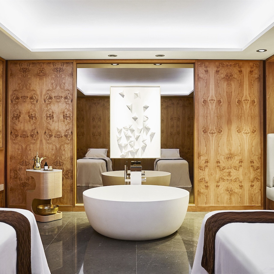 A hotel room with a round bathtub, two beds, a sofa, a desk with a lamp, and a decorative wall panel.