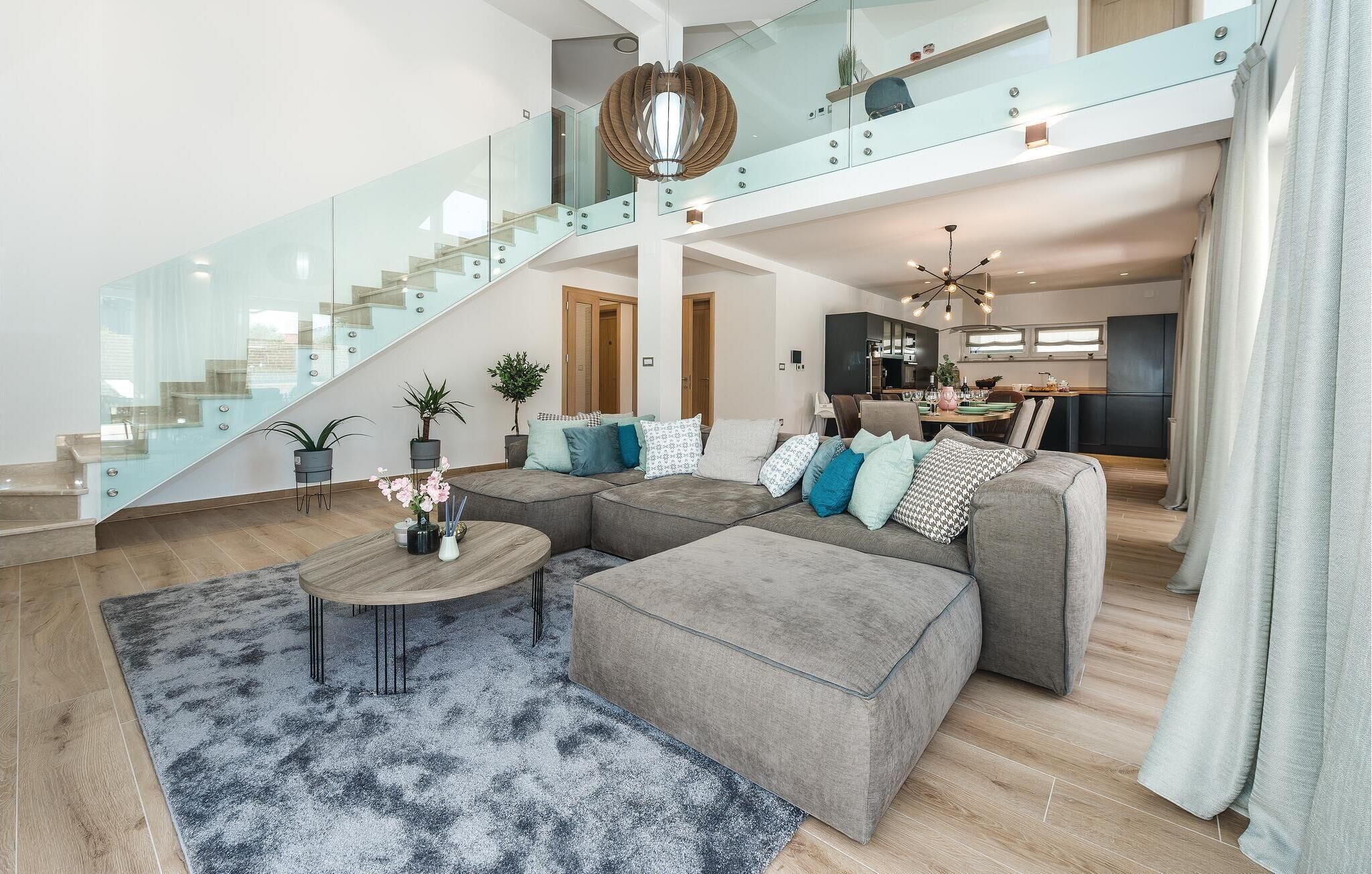 A spacious living area with a large grey sectional sofa, a glass staircase, and a modern kitchen in the background.