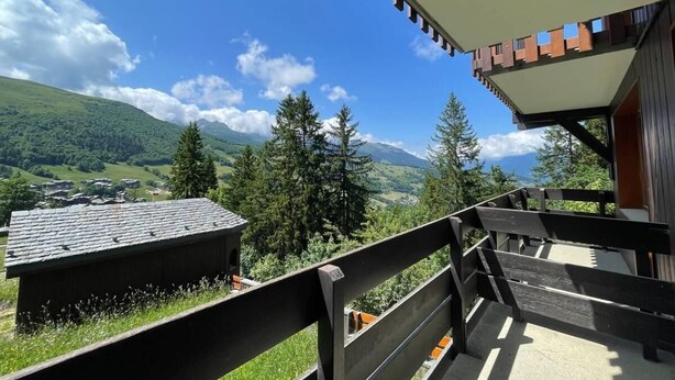 Un balcón con vistas a un paisaje montañoso, terraza de madera y un edificio con tejado inclinado.