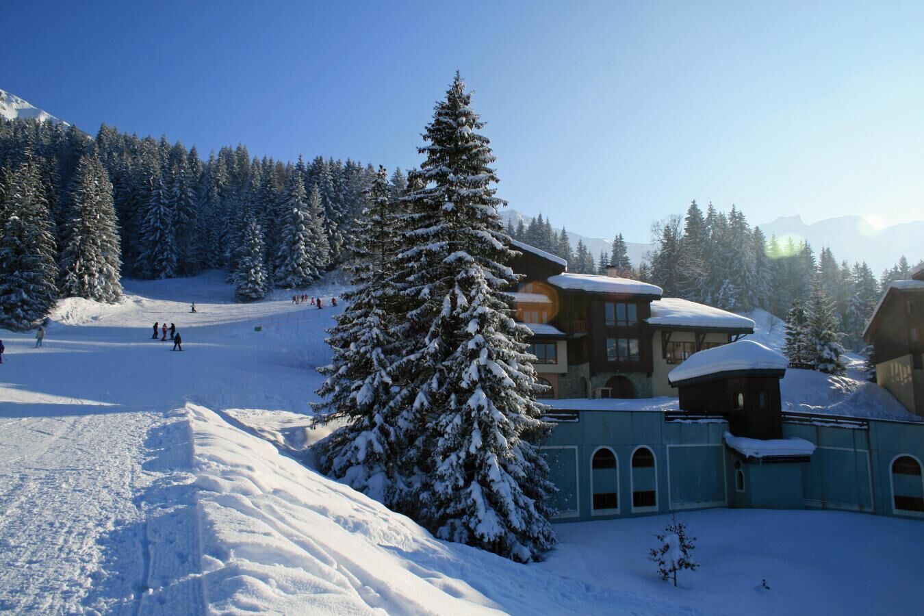 Estación de esquí nevada con un albergue que tiene un árbol grande al frente, una pista de esquí y montañas al fondo.