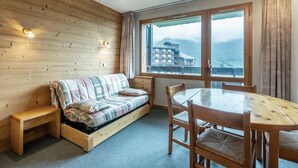 Un cuarto acogedor con pared de madera, un sofá, una mesa de comedor y sillas. Una ventana ofrece vistas a un edificio y montañas.