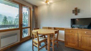 Una habitación acogedora con una mesa y sillas de madera, un televisor sobre un mueble de madera, y un ventanal con vistas a árboles y montañas.