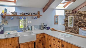 A kitchen with wooden cabinets, a white countertop, and a stone wall. There are hanging pots and pans, a sink, and a window with a view of greenery. - 3 bedroom accommodation in Menheniot, near Liskeard (Menheniot, near Liskeard)