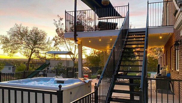 Un jacuzzi con baranda de metal, una escalera que lleva a un espacio al aire libre cubierto, y un edificio de ladrillo al fondo.