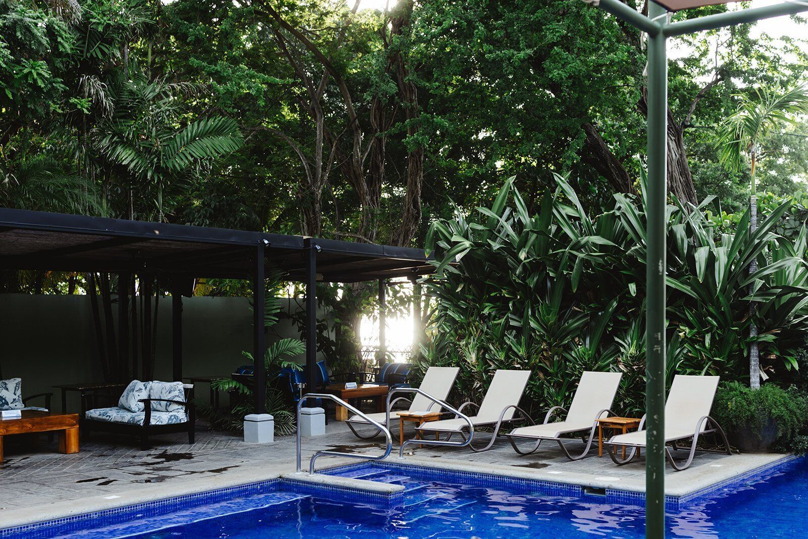 Un espace piscine doté de chaises longues et d’un coin salon abrité, le tout entouré d’une végétation luxuriante.