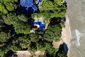 Vue aérienne d’une propriété située sur la côte, avec un bâtiment à toit bleu entouré d’une végétation luxuriante et bordé d’une plage de sable.