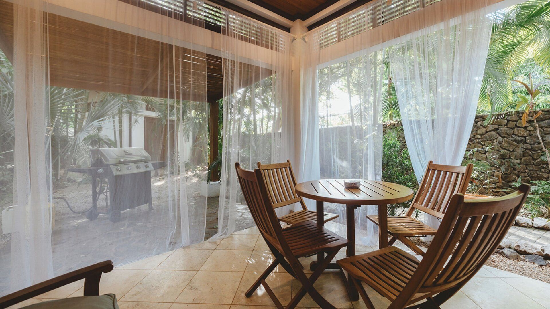 An outdoor patio with a round wooden table and four wooden chairs, surrounded by sheer white curtains and a stone wall.