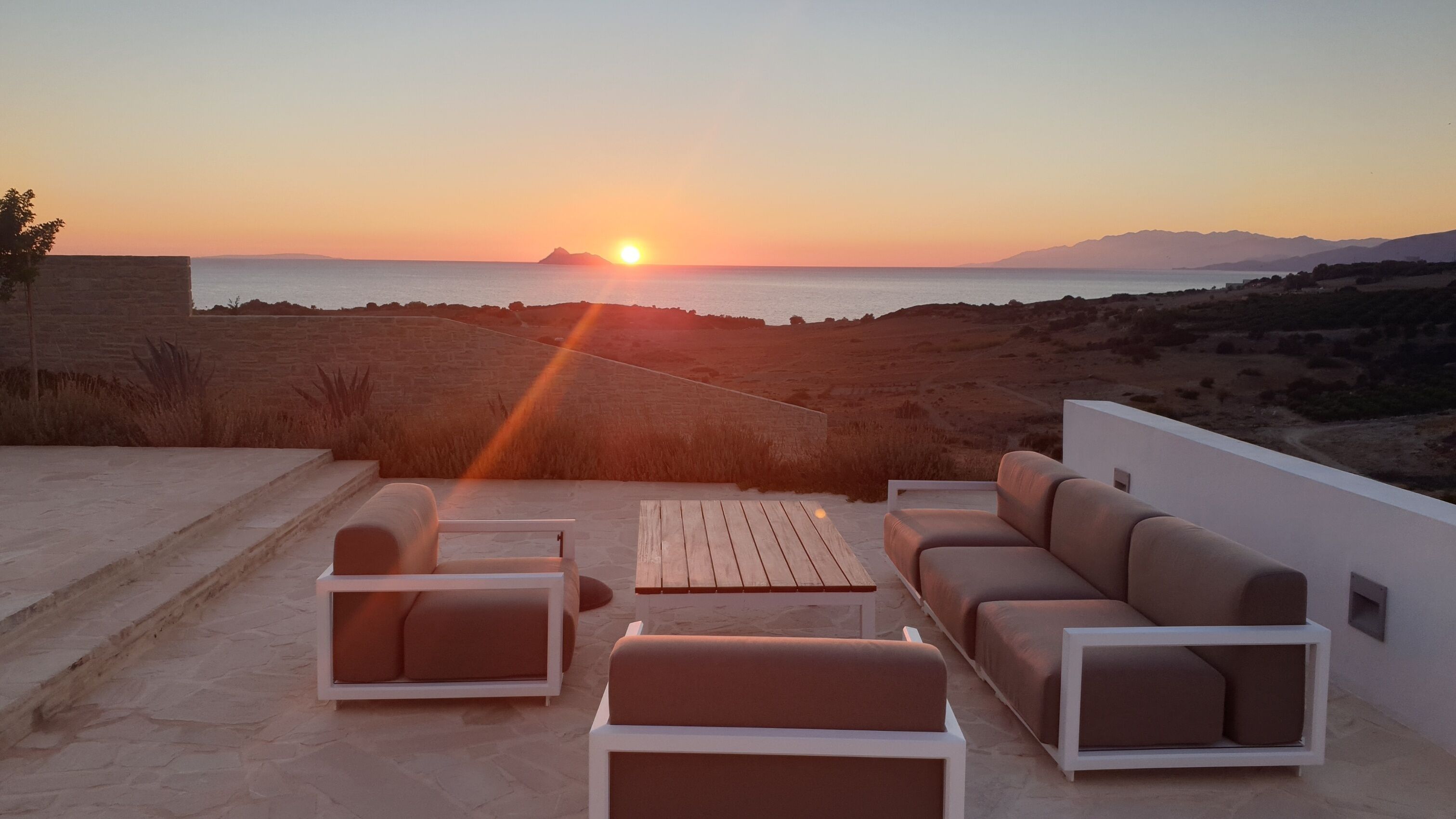 Um terraço com sofá, cadeiras e mesa, com vista para o pôr do sol sobre a paisagem costeira.