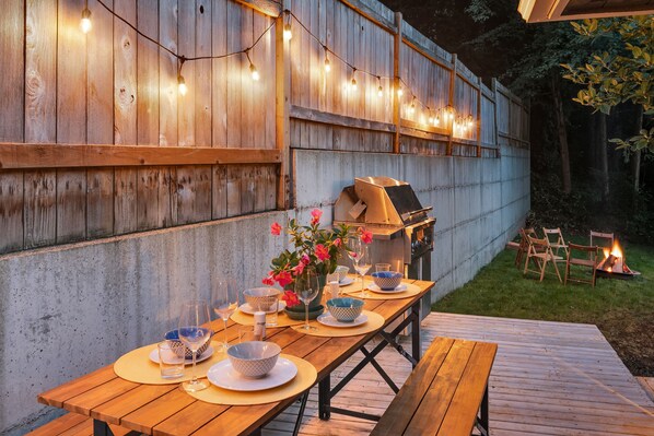An outdoor dining area with a wooden table set for four, a grill, and string lights. - Spacious Main Street Retreat ~ Backyard ~ Parking! (Edmonds)