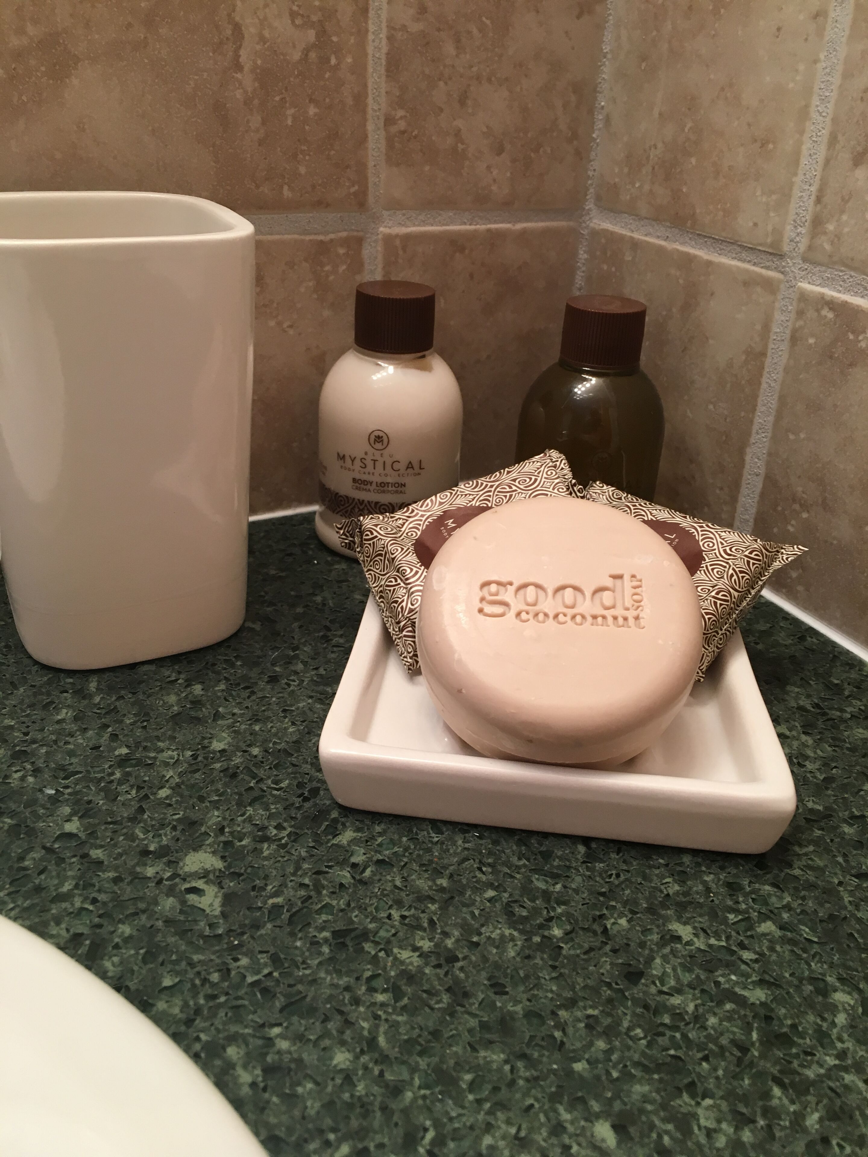 Uma bancada de banheiro com uma caneca branca grande, um frasco de loção corporal Mystical e um sabonete em barra com aroma de coco.