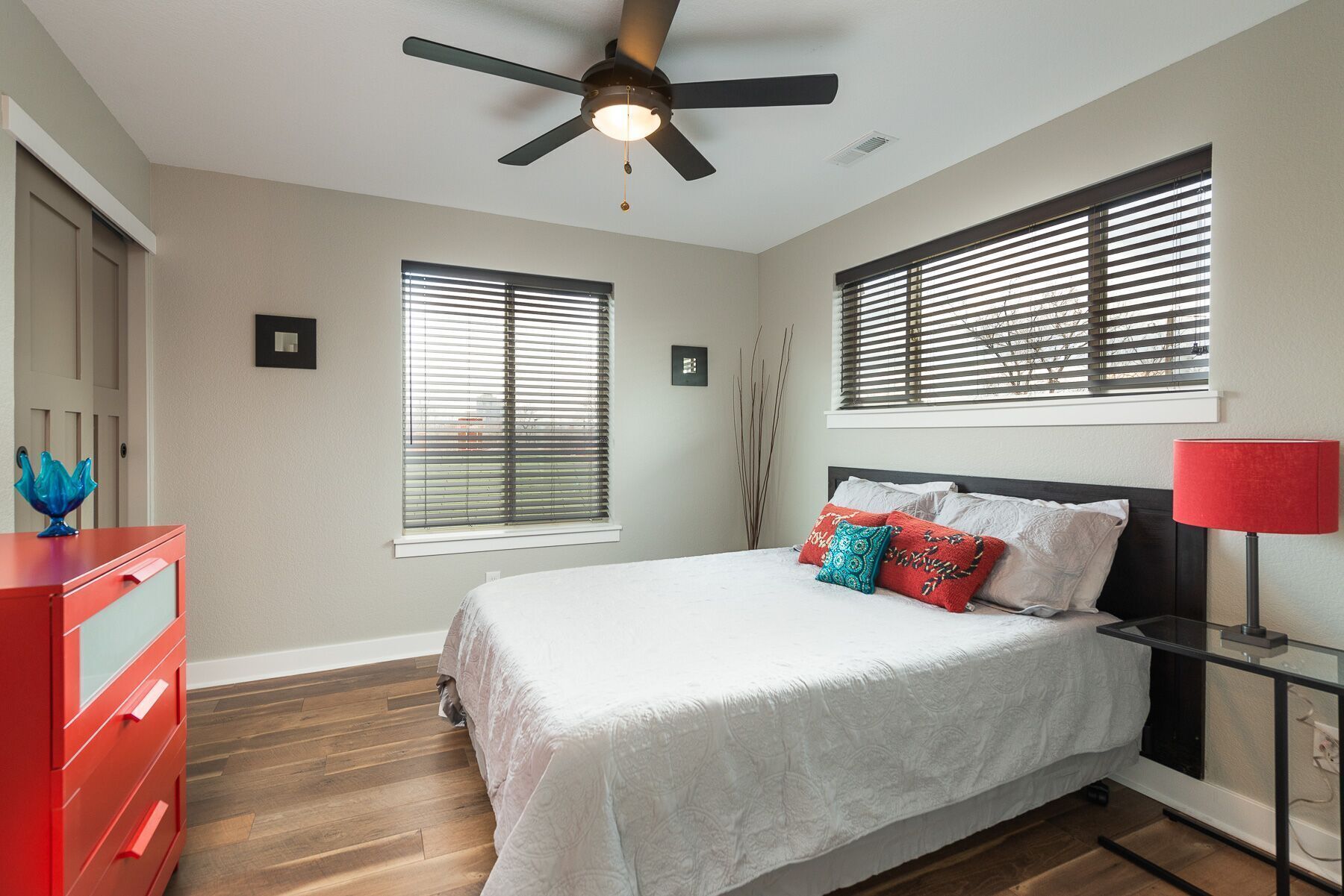 Une chambre à coucher comprenant un lit, un ventilateur de plafond, deux fenêtres avec stores, une commode rouge et une table de chevet avec une lampe.