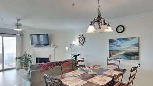 A dining area with a wooden table, chairs, and a chandelier. There is a sofa and a television in the background. - Oceanfront condo w. beach access + balcony views. (Tybee Island)