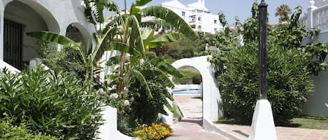 A pathway leading to a white building with arches, surrounded by lush greenery and a lamp post.