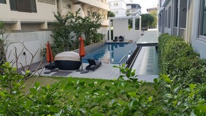 A modern outdoor pool area with lounge chairs and umbrellas, surrounded by greenery and adjacent to a building.
