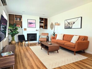 A living room with a fireplace, a sofa, a coffee table, a bookshelf, and a large plant. - Private stargazing retreat with Firepit/Views/ Spa/King bed (Joshua Tree)