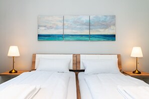 A hotel room with two beds, white linens, a wooden headboard, and a large painting of a beach scene above the beds. - Mia - Exclusive vacation home with sauna, fireplace, 4 pers. (Vitzdorf)