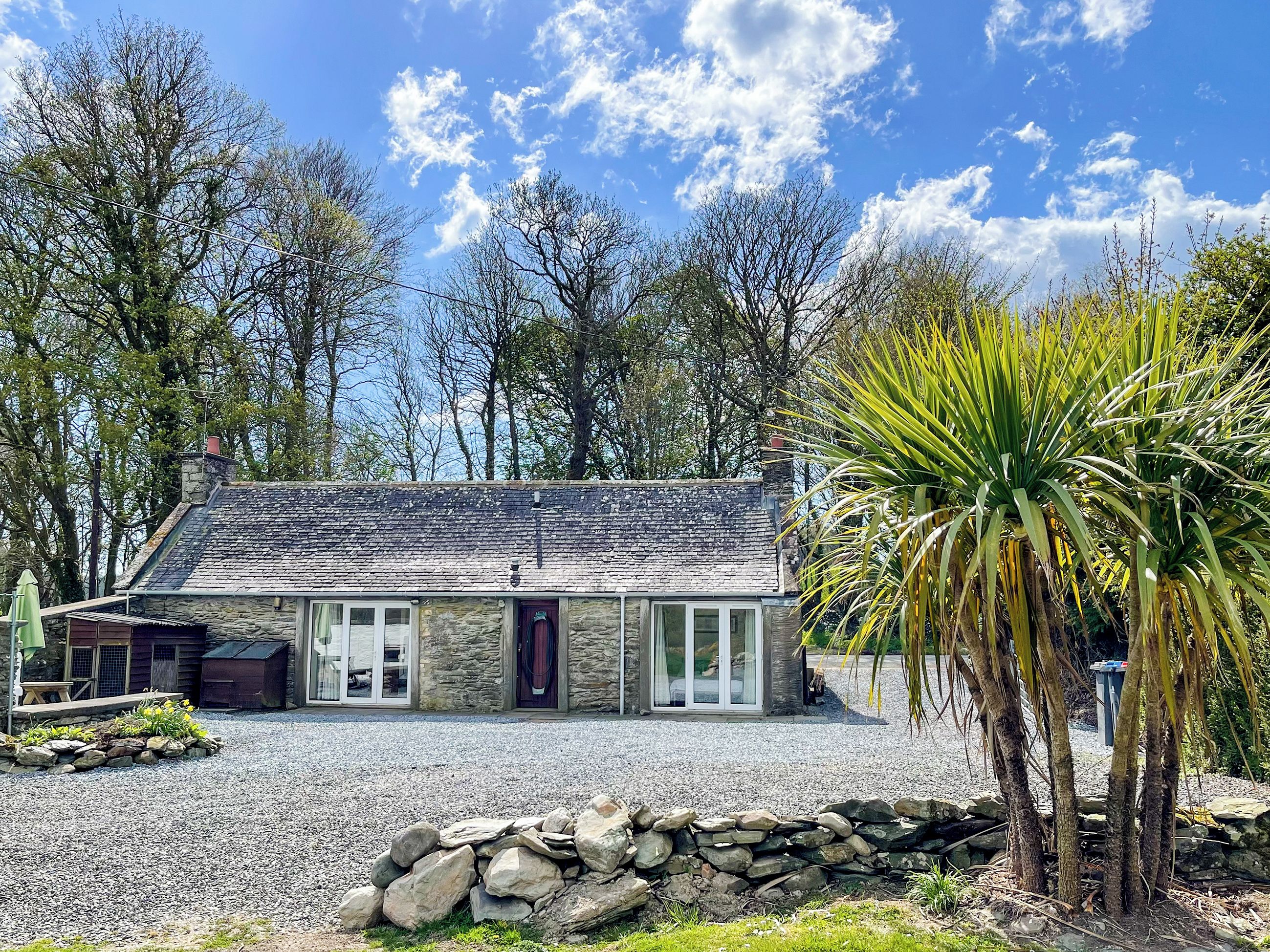 A stone cottage with a gravel driveway, a stone wall, and a palm tree.