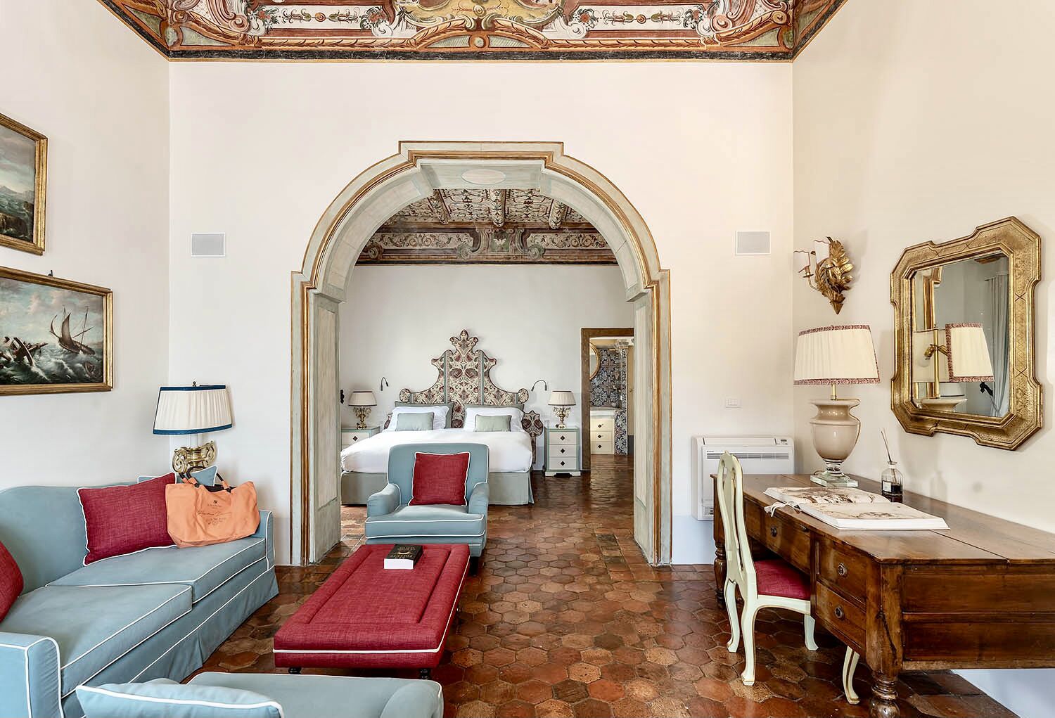 A hotel room with a large bed, a sitting area with a sofa and armchairs, a desk with a chair, and a decorative ceiling.