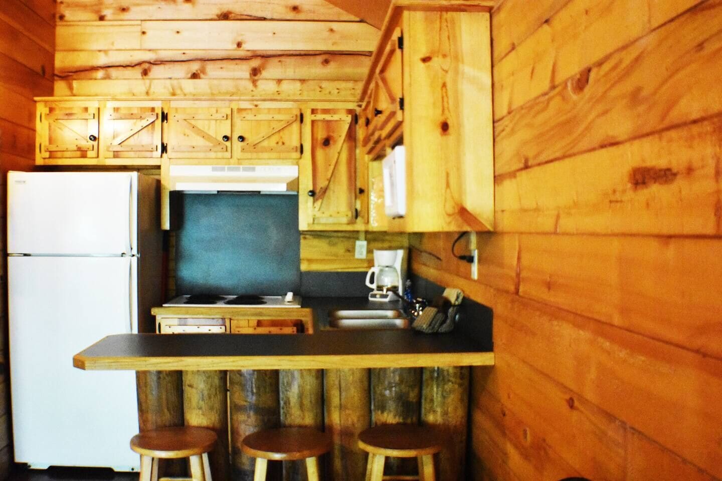 Une cuisine de taille réduite, dotée de meubles en bois, d’un réfrigérateur, d’une cuisinière et de tabourets de bar.