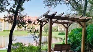 Uma pérgola de madeira com um banco, rodeada por vegetação exuberante e um lago.