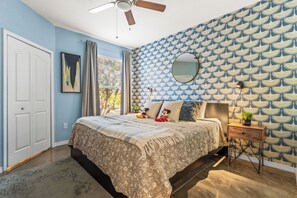 Un dormitorio con cama, ventilador de techo, mesita de noche con lámpara y ventana con cortinas.