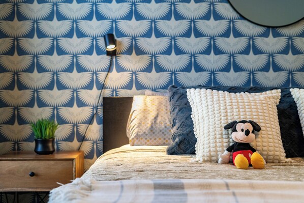 Una cama bien hecha con un suave juguete de Mickey Mouse, una lámpara de noche y un papel tapiz estampado.