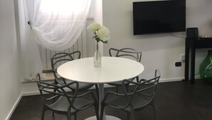 Uma sala de jantar com uma mesa redonda, seis cadeiras, um vaso com flores brancas, uma televisão na parede e uma janela com cortinas leves.