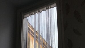 Uma janela com persianas verticais parcialmente abertas, mostrando a fachada de um edifício com várias janelas e portadas.