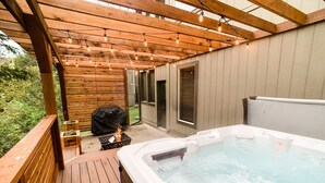 Ein Whirlpool unter einer Holzpergola mit Lichterketten, einem Grill und Blick auf ein Haus.